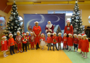 Grupa dzieci stoi z panią dyrektor Marią Królikowską i panią Eweliną Cichą, w tle dekoracja świąteczna, choinki.